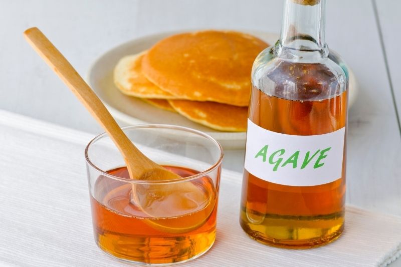 Syrop z agawy w szklanej butelce, obok produkt w szklance, w tle znajdują się amerykańskie naleśniki 