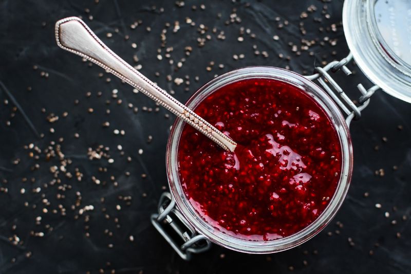 Zbliżenie na słoik pełny dżemu truskawkowego z nasionami chia, na czarnym tle rozsypane nasionka