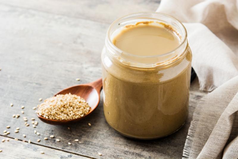 Pasta tahini w słoiku, obok ziarna sezamu na drewnianej łyżce